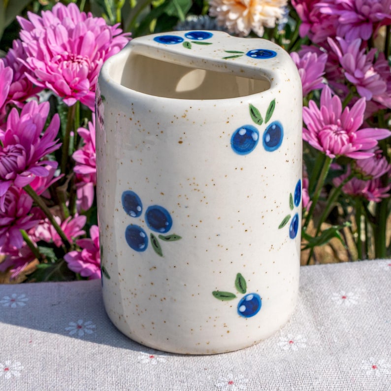 May include: A ceramic toothbrush holder with a cream-colored speckled finish. The holder is decorated with hand-painted blue blueberries and green leaves. The holder has a curved top with a slot for toothbrushes.