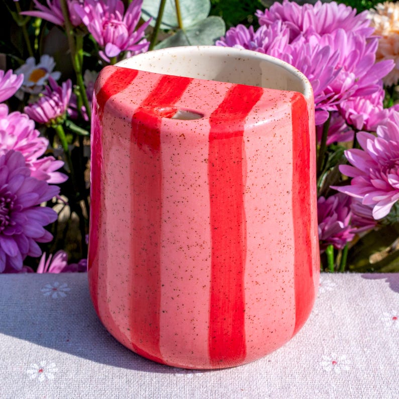 Peut inclure: Un gobelet en c&eacute;ramique avec une finition mouchet&eacute;e et des rayures verticales altern&eacute;es rouge vif et rose tendre. Le r&eacute;cipient pr&eacute;sente une large ouverture sup&eacute;rieure avec un int&eacute;rieur cr&egrave;me lisse et une petite ouverture circulaire pr&egrave;s du bord.