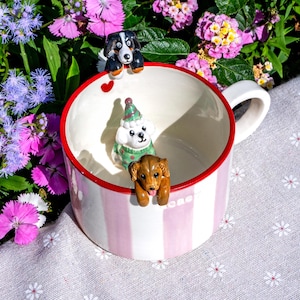 May include: A striped ceramic mug with a red rim, decorated with three dog figurines. The dogs are black and white, white with a green sweater, and brown. The mug is set against a backdrop of colorful flowers. The mug is white with pink stripes.
