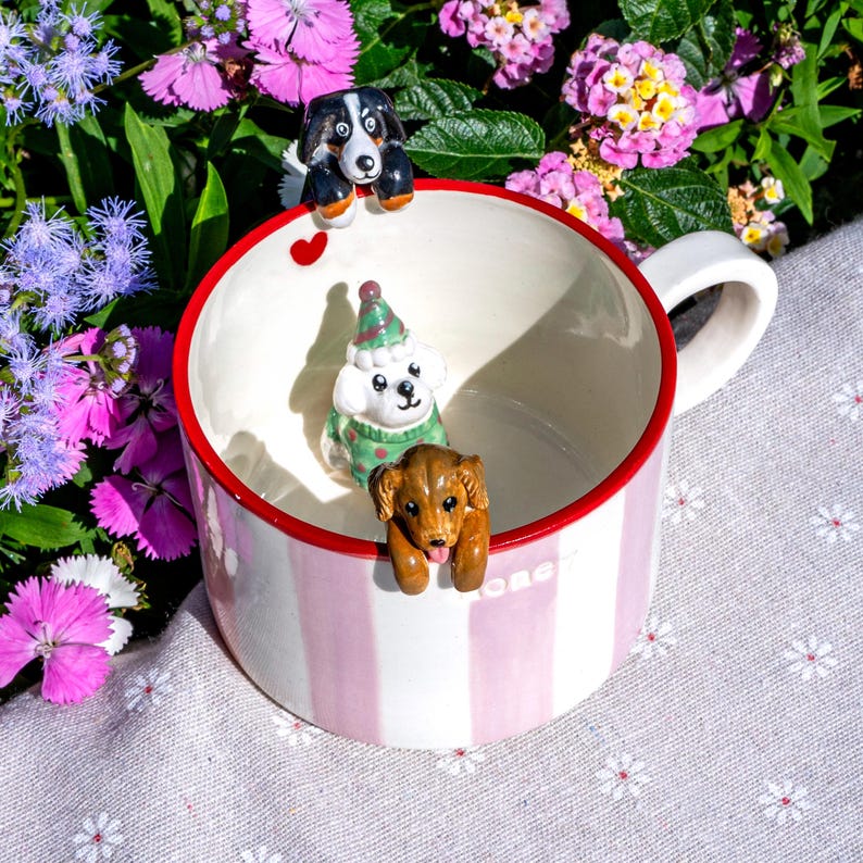 May include: A striped ceramic mug with a red rim, decorated with three dog figurines. The dogs are black and white, white with a green sweater, and brown. The mug is set against a backdrop of colorful flowers. The mug is white with pink stripes.