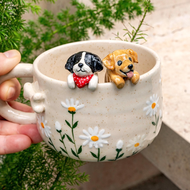 Puede incluir: Una taza de cer&aacute;mica color crema con acabado moteado, decorada con margaritas blancas pintadas a mano y tallos verdes. Dos figuras de perro esculpidas, una negra y blanca con un pa&ntilde;uelo rojo de lunares y otra de color marr&oacute;n dorado, asoman por el borde.