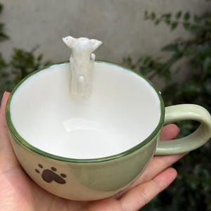 May include: A light green ceramic mug with a white interior and a small white dog figurine attached to the inner rim. The exterior features a dark brown paw print design, a smooth finish, and a curved handle on the side of the round vessel.