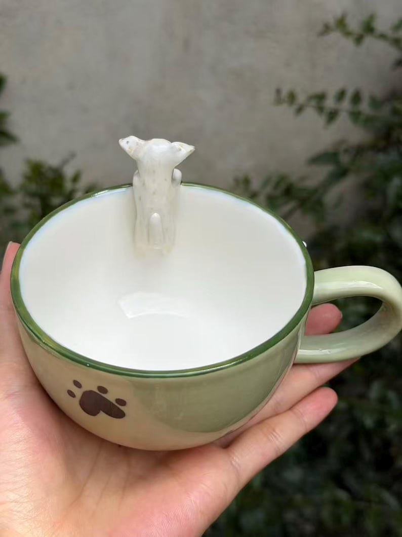 May include: A light green ceramic mug with a white interior and a small white dog figurine attached to the inner rim. The exterior features a dark brown paw print design, a smooth finish, and a curved handle on the side of the round vessel.