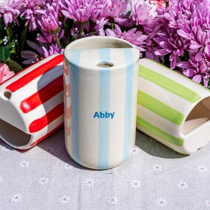 May include: Three cylindrical ceramic toothbrush holders with vertical stripes in red, light blue, and light green. The centre holder features the name Abby printed in blue. Each holder has a circular opening at the top and a large side cutout.