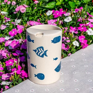 May include: A cylindrical, off-white travel mug with a dark blue fish pattern. The mug has a small opening on the top for drinking. The background features vibrant pink and white flowers, suggesting an outdoor setting.