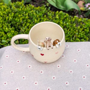 K&ouml;nnte beinhalten: Eine wei&szlig;e Keramiktasse mit rosa Herzen und drei kleinen Hundefiguren, die &uuml;ber den Rand schauen. Die Tasse hat einen Henkel und steht auf einem wei&szlig;en Stoff mit Blumenmuster.