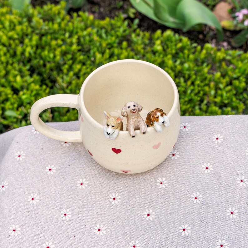 K&ouml;nnte beinhalten: Eine wei&szlig;e Keramiktasse mit rosa Herzen und drei kleinen Hundefiguren, die &uuml;ber den Rand schauen. Die Tasse hat einen Henkel und steht auf einem wei&szlig;en Stoff mit Blumenmuster.