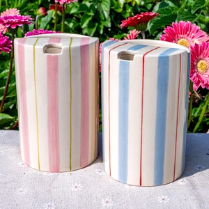May include: Two semi-cylindrical ceramic containers featuring vertical stripes. The left container has pink and yellow stripes on a cream background, while the right container has blue and red stripes on a cream background. Both have a top opening.