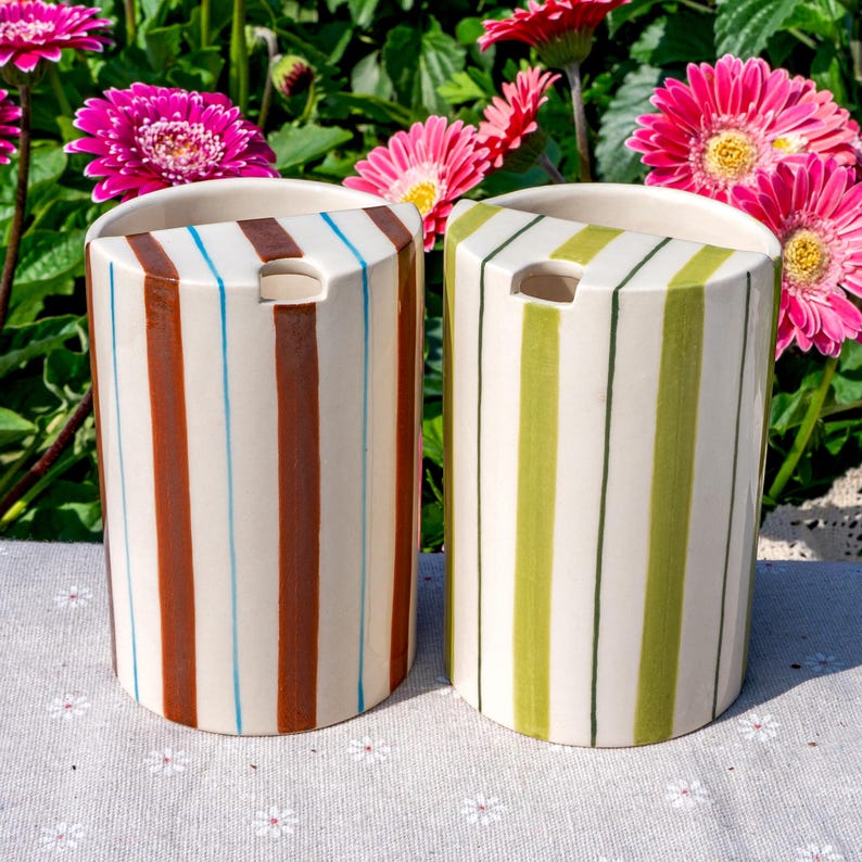 May include: Two semi-cylindrical ceramic containers featuring vertical stripes. The left container has brown and light blue stripes on a cream base. The right container has light and dark green stripes on a cream base. Both have a top opening.