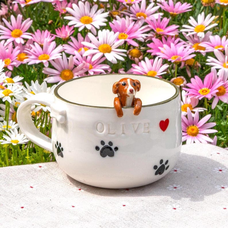 May include: White ceramic mug with the name "OLIVE", a red heart, and black paw prints. A small brown and white dog figurine is perched on the rim. The mug is set against a backdrop of pink and white daisies.