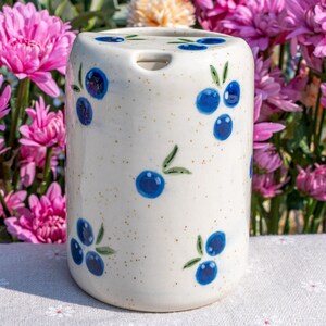 May include: A cylindrical ceramic toothbrush holder with a speckled cream background. The holder is decorated with hand-painted blue blueberries and green leaves. The holder has a small opening at the top for a toothbrush.