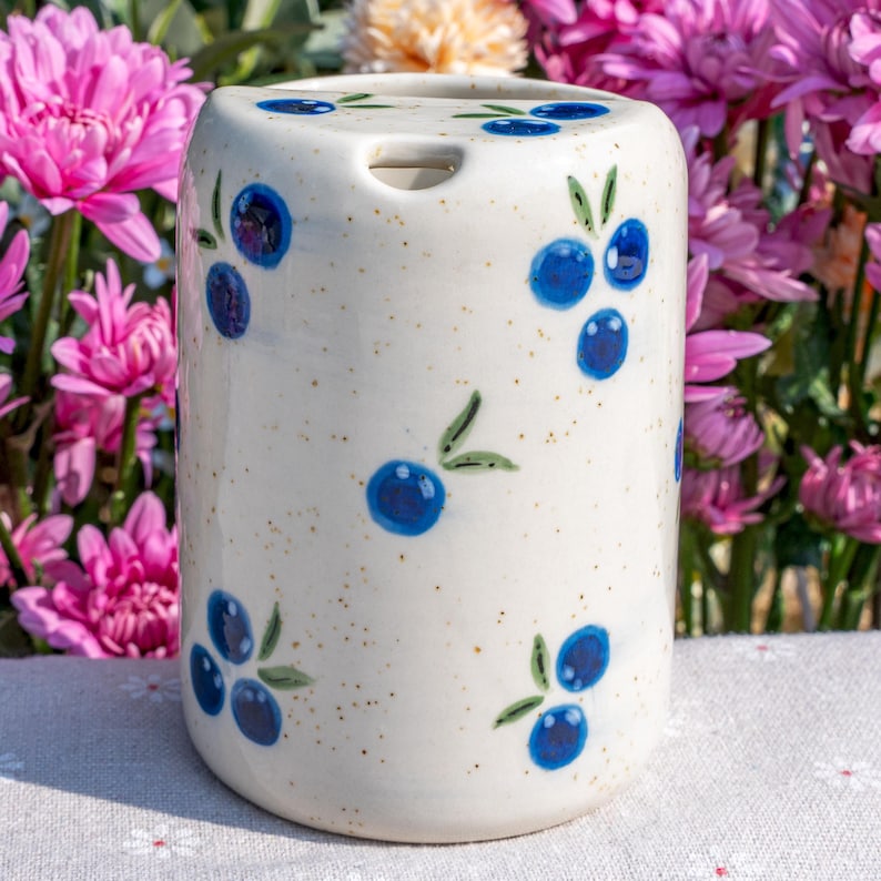 May include: A cylindrical ceramic toothbrush holder with a speckled cream background. The holder is decorated with hand-painted blue blueberries and green leaves. The holder has a small opening at the top for a toothbrush.