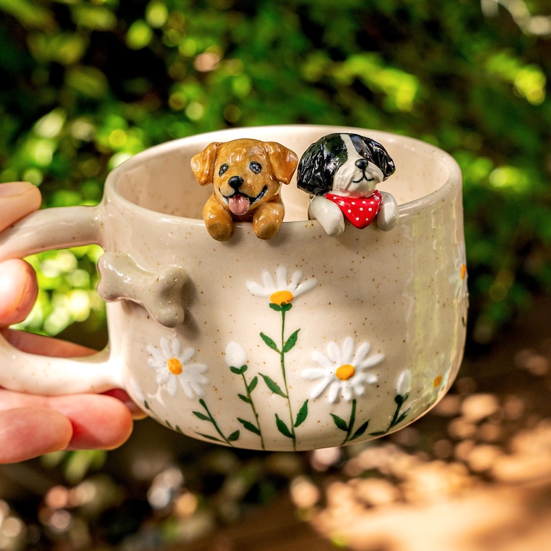 Puede incluir: Una taza de cer&aacute;mica color crema con acabado moteado y margaritas blancas pintadas con tallos verdes. Un hueso esculpido est&aacute; unido al asa, mientras que dos peque&ntilde;as figuras de perros, una dorada y otra blanca y negra, asoman por el borde.