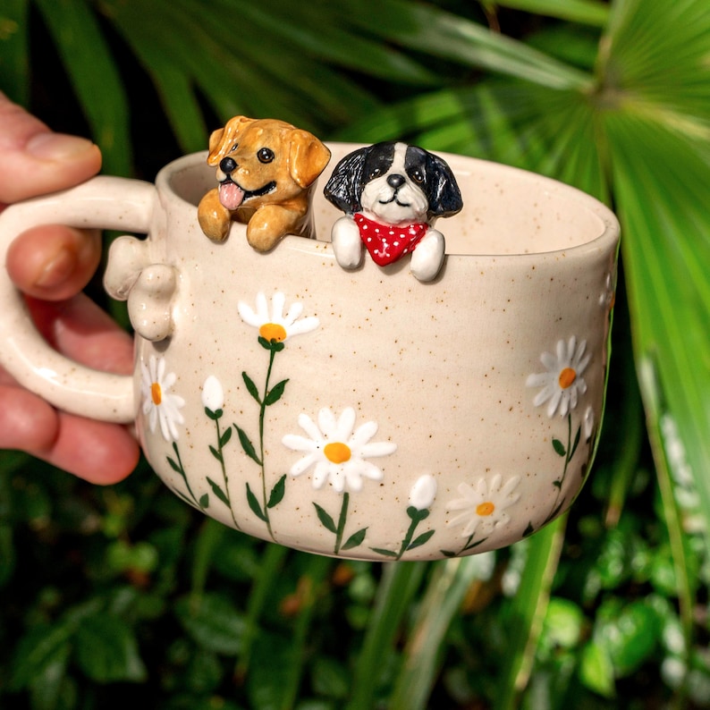 Puede incluir: Una taza de cer&aacute;mica beige con acabado moteado, decorada con margaritas blancas pintadas a mano con centros amarillos. Dos figuras de perro esculpidas est&aacute;n fijadas al borde interior: un perro marr&oacute;n dorado y un perro blanco y negro con pa&ntilde;uelo.
