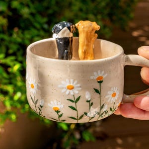 Puede incluir: Una taza de cer&aacute;mica beige con acabado moteado y un patr&oacute;n pintado de margaritas blancas con centros amarillos y tallos verdes. Dentro de la taza, dos peque&ntilde;as figuras de perros esculpidas, una negra con blanco y otra color canela, est&aacute;n de pie.