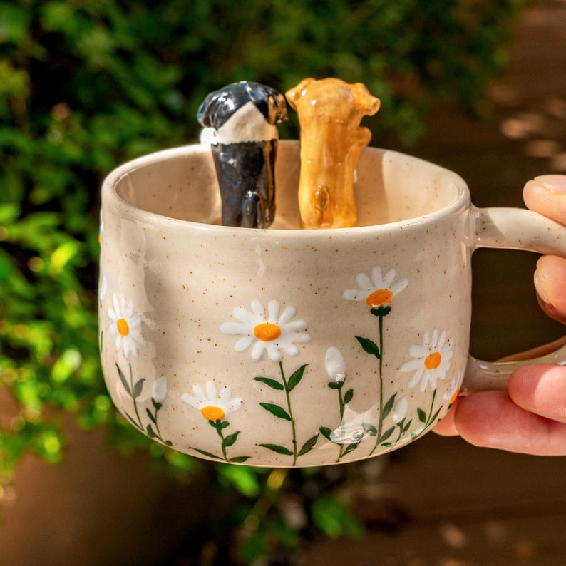 Puede incluir: Una taza de cer&aacute;mica beige con acabado moteado y un patr&oacute;n pintado de margaritas blancas con centros amarillos y tallos verdes. Dentro de la taza, dos peque&ntilde;as figuras de perros esculpidas, una negra con blanco y otra color canela, est&aacute;n de pie.