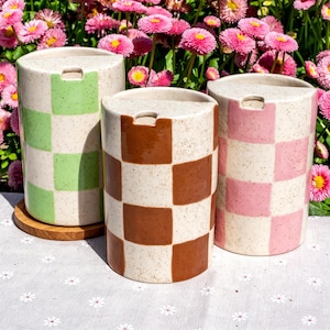 May include: Three cylindrical ceramic tumblers with a speckled cream base and a checkered pattern. One tumbler features light green squares, one features brown squares, and one features pink squares. Each tumbler has a flat top with a small notch.