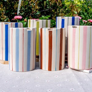 May include: A collection of five cylindrical ceramic containers, each featuring vertical stripes in various colors including blue, light blue, green, brown, and pink. Each vessel has a smooth, glossy finish and a small notch at the top rim.