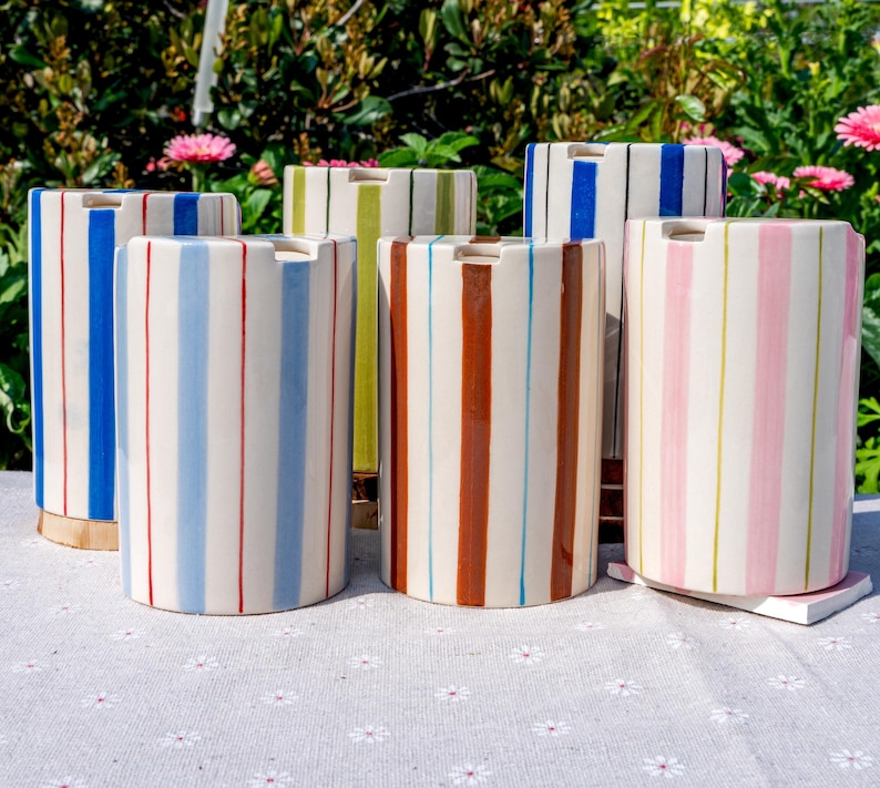 May include: A collection of five cylindrical ceramic containers, each featuring vertical stripes in various colors including blue, light blue, green, brown, and pink. Each vessel has a smooth, glossy finish and a small notch at the top rim.