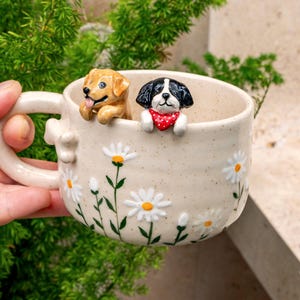 Puede incluir: Una taza de cer&aacute;mica color crema con acabado moteado, decorada con margaritas blancas pintadas a mano con centros amarillos. Dos peque&ntilde;as figuras de perros esculpidas, una dorada y otra negra y blanca con un pa&ntilde;uelo rojo de lunares, asoman por el borde.