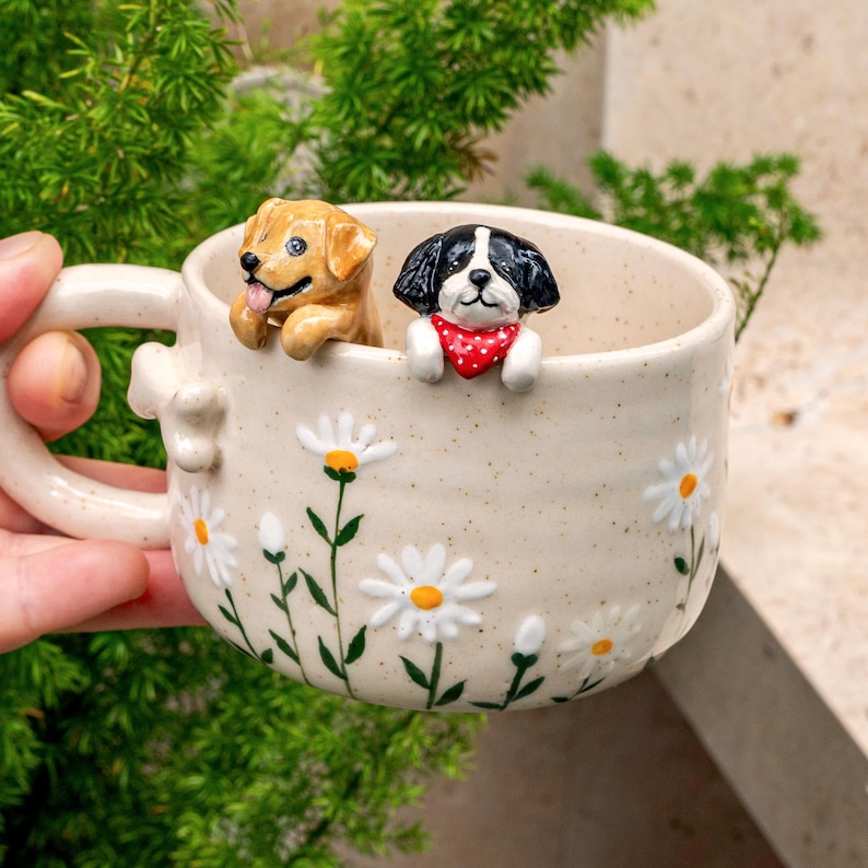 Puede incluir: Una taza de cer&aacute;mica color crema con acabado moteado, decorada con margaritas blancas pintadas a mano con centros amarillos. Dos peque&ntilde;as figuras de perros esculpidas, una dorada y otra negra y blanca con un pa&ntilde;uelo rojo de lunares, asoman por el borde.