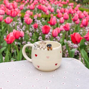 K&ouml;nnte beinhalten: Eine wei&szlig;e Keramiktasse mit pinkfarbenen und roten Herzen, die darauf gedruckt sind. Drei kleine Keramik-Hundefiguren schauen &uuml;ber den Rand der Tasse. Die Tasse steht auf einer wei&szlig;en Tischdecke mit Blumenmuster.