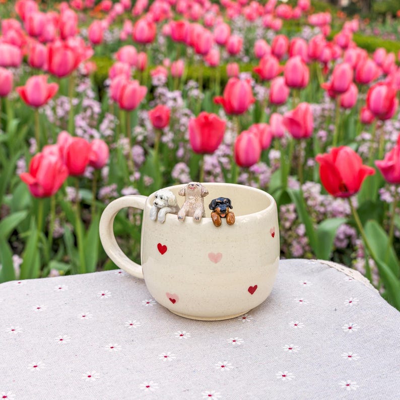 K&ouml;nnte beinhalten: Eine wei&szlig;e Keramiktasse mit pinkfarbenen und roten Herzen, die darauf gedruckt sind. Drei kleine Keramik-Hundefiguren schauen &uuml;ber den Rand der Tasse. Die Tasse steht auf einer wei&szlig;en Tischdecke mit Blumenmuster.
