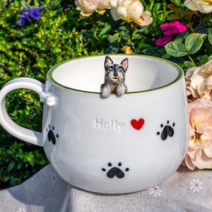 May include: A white ceramic mug with a green rim, featuring a small sculpted dog figurine peering over the edge. The exterior is decorated with black paw prints, a red heart, and the embossed name Holly. A small bone detail sits on the handle.