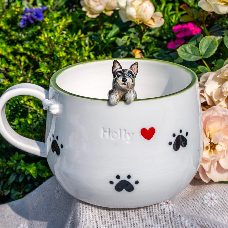 May include: A white ceramic mug with a green rim, featuring a small sculpted dog figurine peering over the edge. The exterior is decorated with black paw prints, a red heart, and the embossed name Holly. A small bone detail sits on the handle.