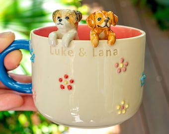 May include: A cream-colored ceramic mug with a blue handle and a coral interior. Two sculpted dog figurines, one white and brown and one golden brown, peek over the rim. The text Luke & Lana is embossed on the front, surrounded by small, raised, multi-colored flower appliques.
