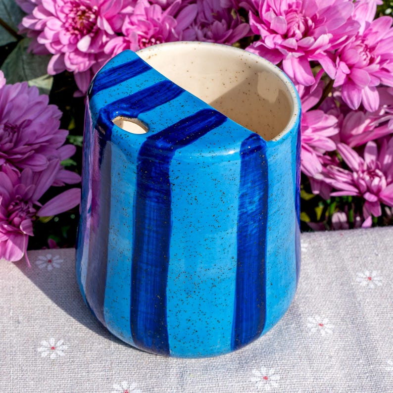 Peut inclure: Une tasse en c&eacute;ramique avec un int&eacute;rieur cr&egrave;me mouchet&eacute; et un ext&eacute;rieur bleu vif orn&eacute; de rayures verticales bleu fonc&eacute;. Le bord pr&eacute;sente une d&eacute;coupe inclin&eacute;e unique avec une petite ouverture circulaire pr&egrave;s du sommet de la paroi.