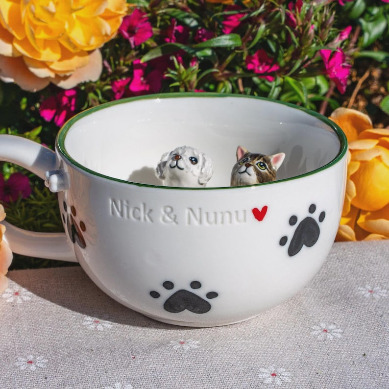 May include: A white ceramic mug with a green rim, featuring black paw prints and the embossed text "Nick & Nunu" with a red heart. Inside the mug, two small 3D animal figurines are visible: a white curly-haired dog and a brown tabby cat.