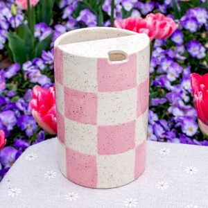 May include: A cylindrical ceramic container featuring a speckled cream and pink checkerboard pattern. The top edge has a flat, semi-circular cutout section and a small, rectangular opening on the side, creating a distinct, functional design.