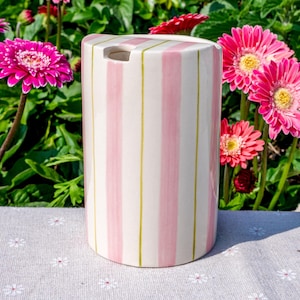May include: A cylindrical ceramic container featuring vertical stripes in alternating shades of soft pink and white, accented by thin, muted green lines. The top surface is flat with a small, circular opening positioned near the edge.