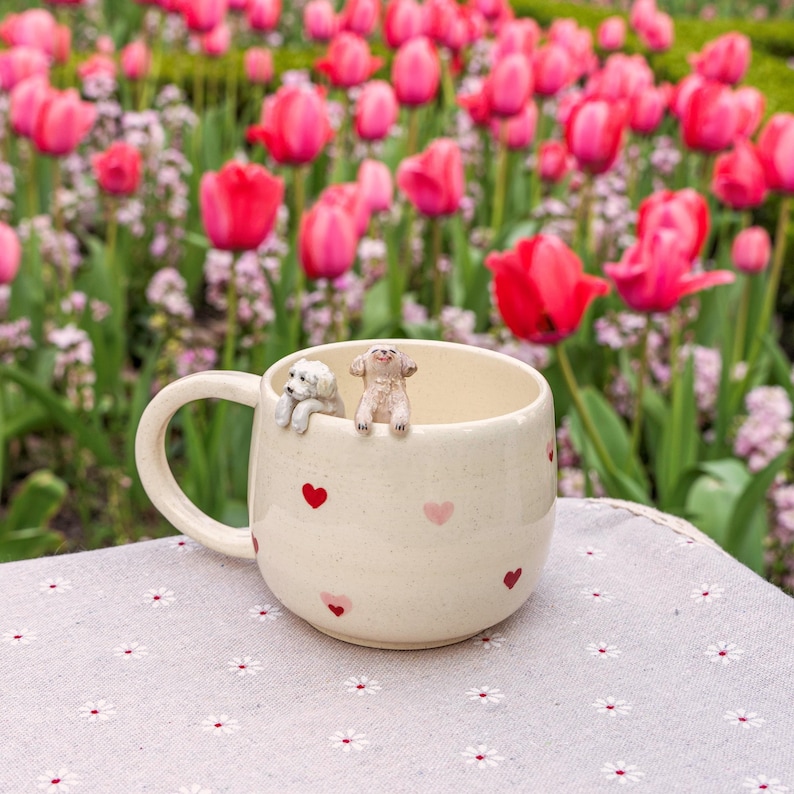 K&ouml;nnte beinhalten: Eine wei&szlig;e Keramiktasse mit pinkfarbenen Herzen bedruckt. Zwei kleine Keramik-Hundefiguren sitzen auf dem Rand der Tasse.