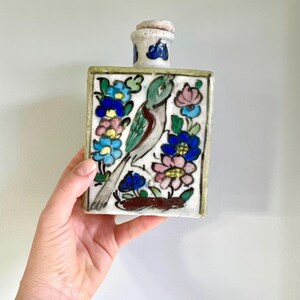 Peut inclure: Une bouteille en céramique rectangulaire à base blanche, ornée d'un motif peint à la main représentant un oiseau entouré de fleurs bleues, roses et vertes. La bouteille possède un col cylindrique court avec un bouchon assorti et une bordure épaisse.