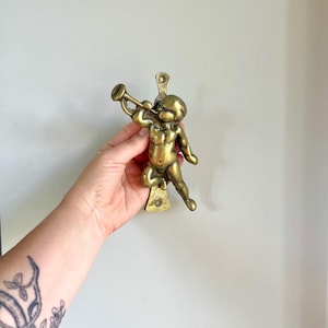 Könnte beinhalten: Ein Türklopfer aus Messing mit einem Posaune spielenden Putto. Der Putto steht, die Trompete an den Mund gehalten. Der Klopfer hat einen warmen Goldton und ist auf einer weißen Oberfläche montiert.