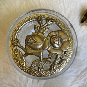 May include: A glass jar with a brass lid featuring a decorative design of two roses and leaves.