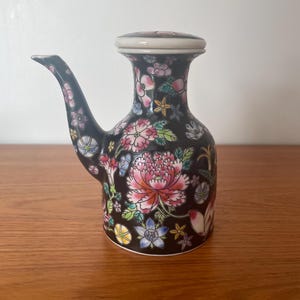 May include: A black ceramic teapot with a floral design. The teapot has a white lid and a spout. The flowers are pink, blue, yellow, and green.