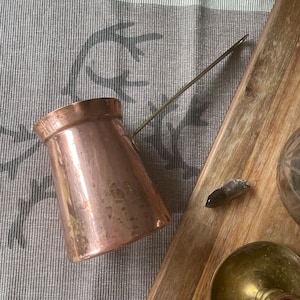 May include: A copper coffee pot with a long, thin handle. The pot is sitting on a grey and white patterned placemat. The pot has a worn, aged look.