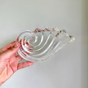 May include: Clear glass decorative dish with a swirling, seashell-like design. The dish has a wave-like shape with a textured surface and a ruffled edge. The glass is transparent, allowing light to pass through, and is held by a hand.