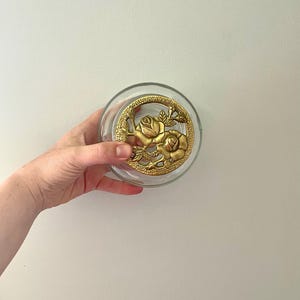 Könnte beinhalten: Eine klare Glasschale mit einem goldfarbenen Blumendesign. Das Design zeigt zwei Rosen und Blätter. Die Schale ist rund und scheint ein dekoratives Stück zu sein.