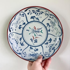 May include: A white ceramic plate with a red rim and blue floral designs. The plate features a central floral motif with green and red accents. The hand-painted design includes stylized flowers and leaves.