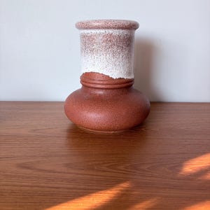 Może przedstawiać: Ceramiczny wazon z szeroką, zaokrągloną podstawą i cylindryczną szyjką. Dolna część ma jednolite, ziemiste wykończenie w kolorze terakoty, podczas gdy górna szyjka pokryta jest nakrapianym, złamany białym szkliwem, które spływa po bokach.