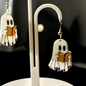 May include: White ghost-shaped earrings with black eyes and a gold book. The earrings are hanging from a gold-colored hook. The ghost has a white body with a purple outline. The book is gold with a brown cover. The earrings are displayed on a white stand.