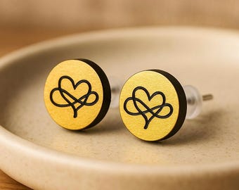 Puede incluir: Un par de pendientes de botón redondos. Cada pendiente presenta una cara dorada con un diseño de corazón infinito negro. Los bordes de los pendientes son negros. Los pendientes se muestran sobre una superficie clara.