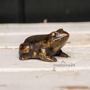 Könnte beinhalten: Eine Bronze-Froschfigur mit detaillierter Textur. Der Frosch sitzt mit ausgestreckten Vorderbeinen und offenen Augen. Die Figur befindet sich auf einer weißen Oberfläche.