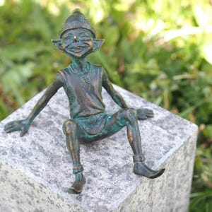 Könnte beinhalten: Bronzestatue eines lächelnden Elfen mit spitzen Ohren und einem konischen Hut. Der Elf sitzt auf einem Steinblock, Arme und Beine ausgestreckt. Die Statue hat eine Grünspan-Oberfläche, was darauf hindeutet, dass sie aus gealtertem Metall besteht.