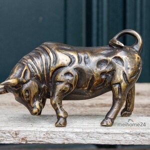 Könnte beinhalten: Bronzefarbene Bullenskulptur mit strukturierter Oberfläche. Der Bulle ist in einer Angriffshaltung dargestellt, mit gesenktem Kopf und Hörnern nach vorne. Die Skulptur steht auf einer Holzoberfläche.