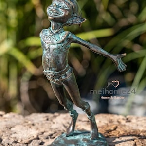 Könnte beinhalten: Eine bronzefarbene Gartenstatue eines lächelnden Elfen mit ausgestreckten Armen. Die Statue hat eine grüne Patina und steht auf einem strukturierten Sockel. Der Elf trägt einen Hut und einen Umhang mit Gürtel. Für Haus- und Gartendekoration.